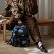 A woman sits on a wooden bench in a minimalist room, wearing a brown bomber jacket and matching pants, with a black backpack featuring a blue and white floral pattern placed in front of her.