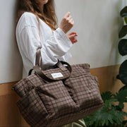 A woman wearing a white long-sleeve shirt leans against a wall while carrying a brown plaid duffel bag with dark brown handles. The bag has a small white label on the front pocket.