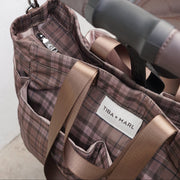 Close-up of a Tiba + Marl stroller bag with a brown and beige plaid pattern, featuring satin brown handles and a white brand label, shown hanging from a stroller handlebar.