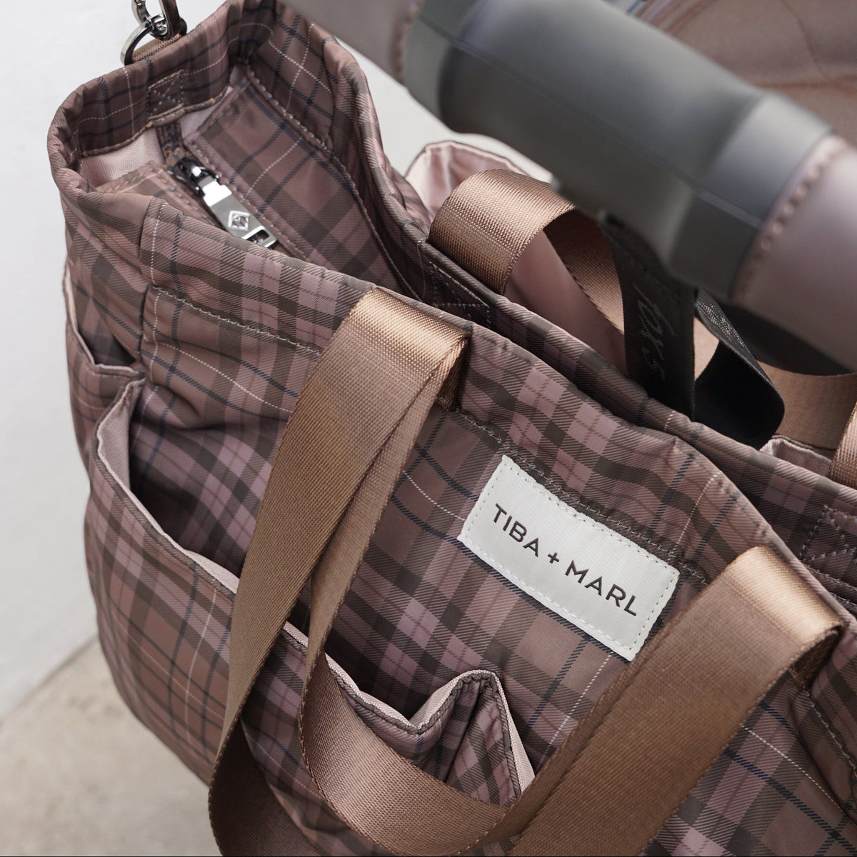 Close-up of a Tiba + Marl stroller bag with a brown and beige plaid pattern, featuring satin brown handles and a white brand label, shown hanging from a stroller handlebar.