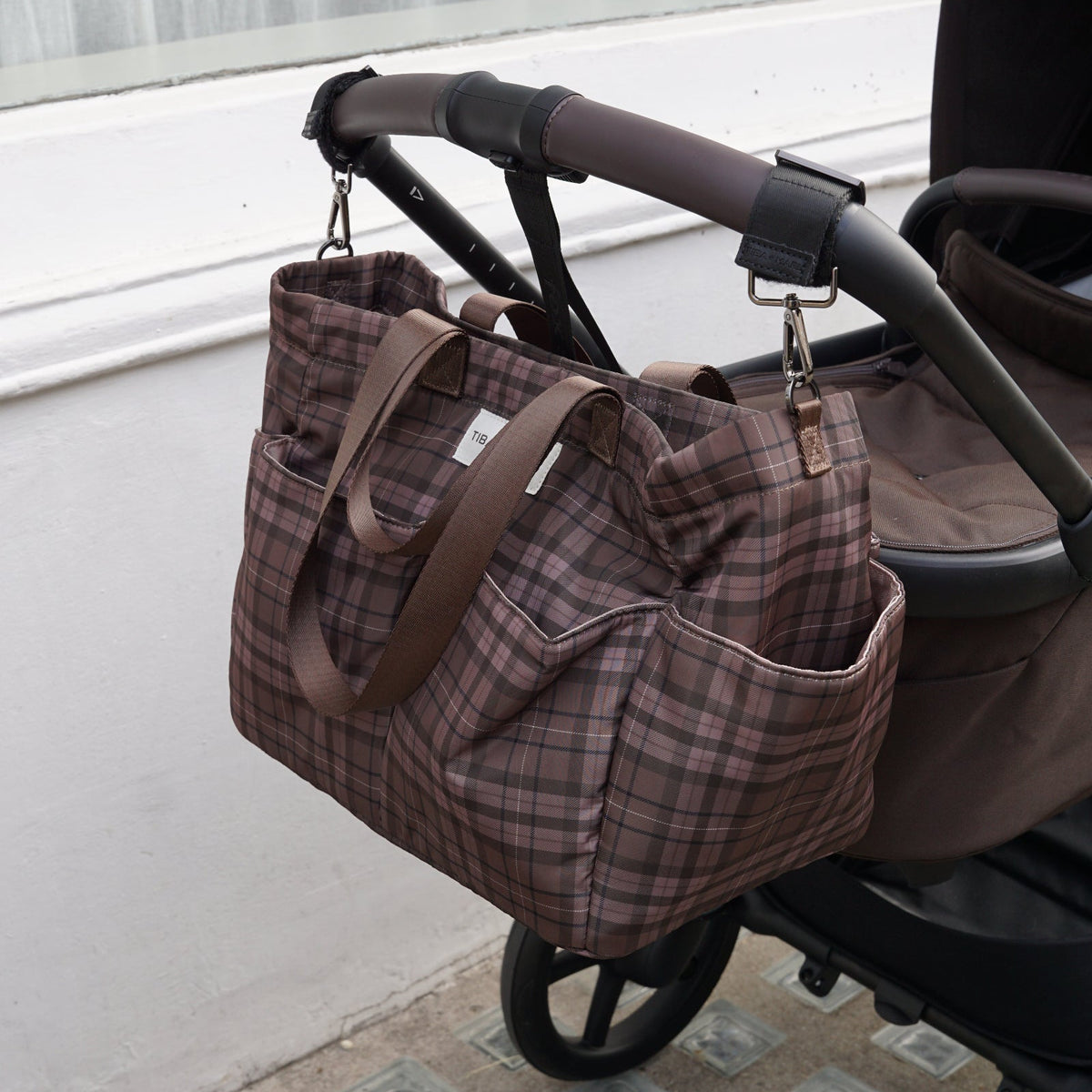 A brown and beige plaid diaper bag with brown leather handles, hanging from a stroller handle outdoors next to a white wall.