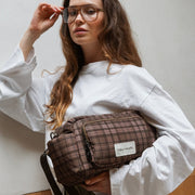 A woman wearing a white sweatshirt and glasses holds a brown plaid crossbody bag with a white brand label that reads 'TIBA + MARL'.