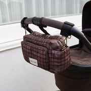 A brown and burgundy plaid diaper bag with a white 'DIANAL' label, hanging from a stroller handlebar in an outdoor setting.