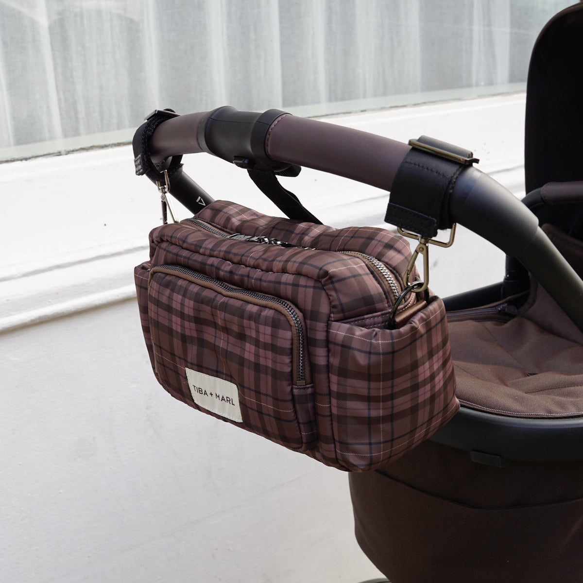 A brown and burgundy plaid diaper bag with a white 'DIANAL' label, hanging from a stroller handlebar in an outdoor setting.
