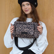 A woman wearing a black New York Yankees beret and a white shirt holds a leopard-print shoulder bag with a black strap against a light wood-paneled background.