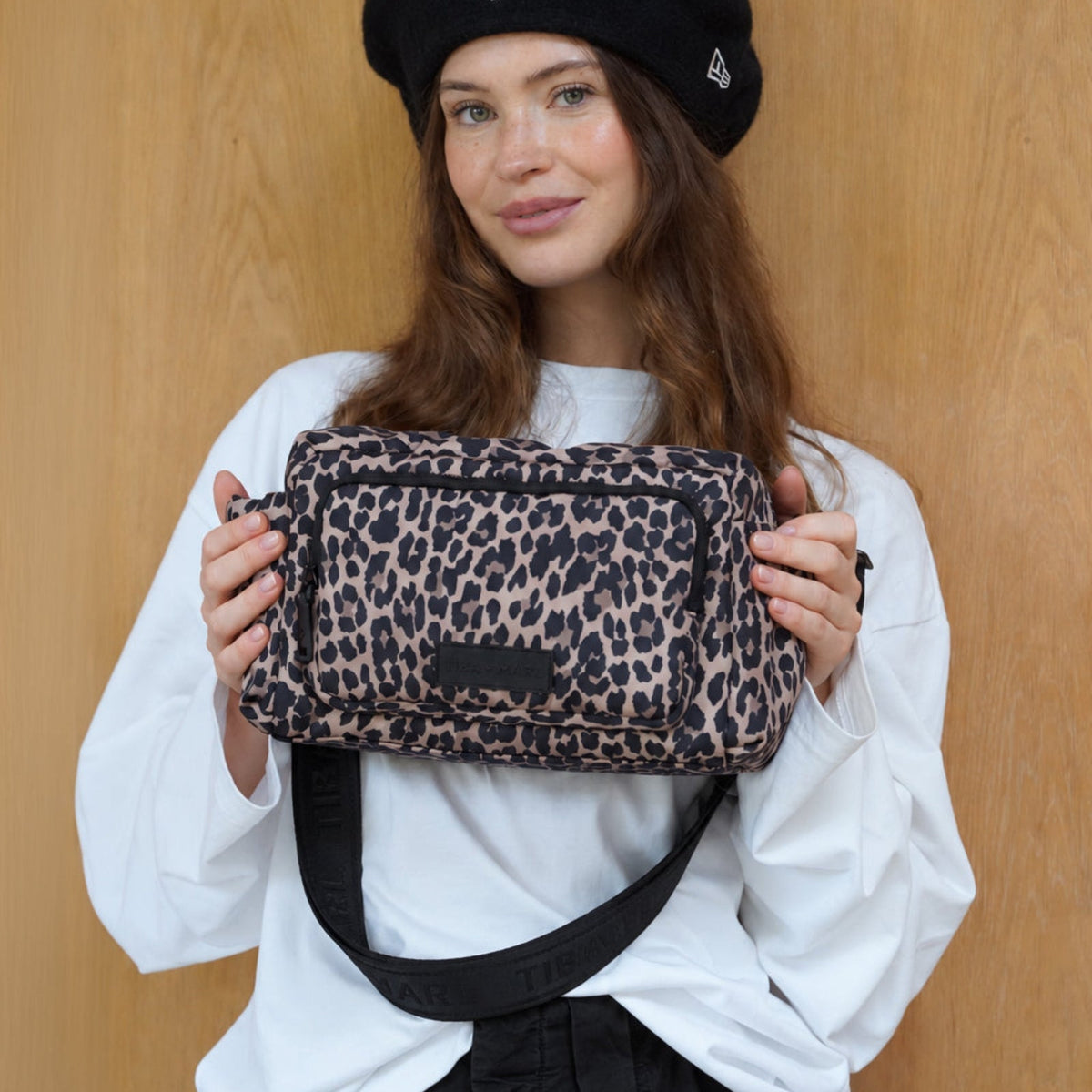 A woman wearing a black New York Yankees beret and a white shirt holds a leopard-print shoulder bag with a black strap against a light wood-paneled background.