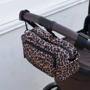 A leopard print diaper bag with black zippers and trim, hanging from the handle of a stroller on an outdoor walkway next to a white wall.