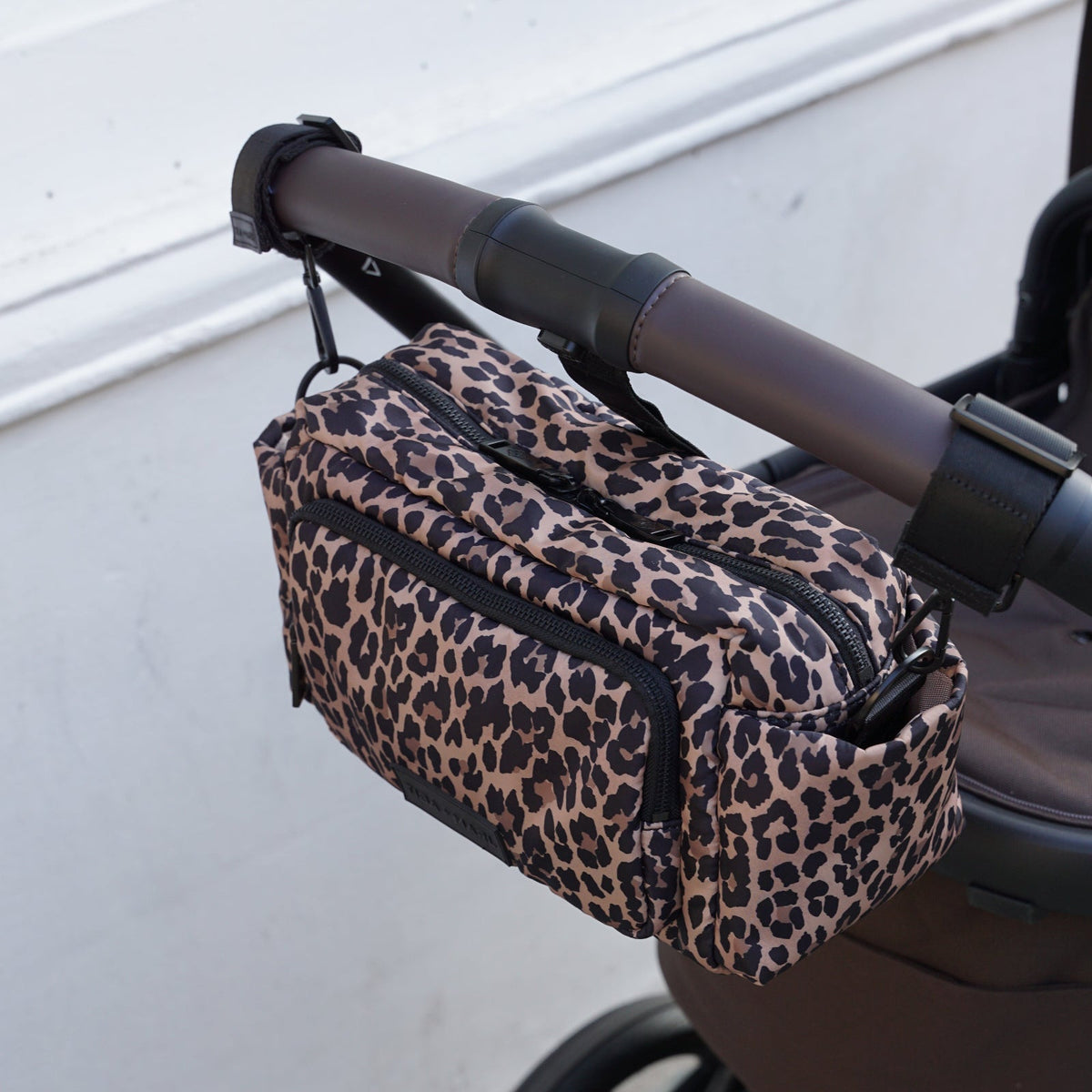 A leopard print diaper bag with black zippers and trim, hanging from the handle of a stroller on an outdoor walkway next to a white wall.