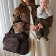A woman holding a baby wears a brown diaper bag over her shoulder. The bag is a dark brown nylon with dual top handles and a front slip pocket, shown in a lifestyle setting against a white curtain background.