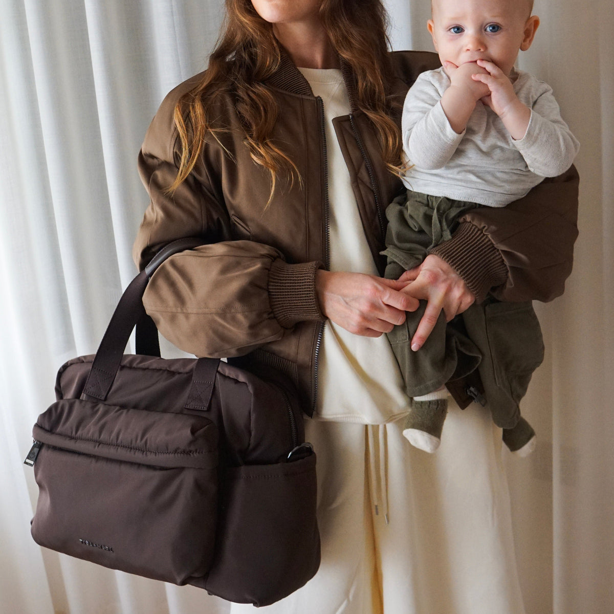 A woman holding a baby wears a brown diaper bag over her shoulder. The bag is a dark brown nylon with dual top handles and a front slip pocket, shown in a lifestyle setting against a white curtain background.