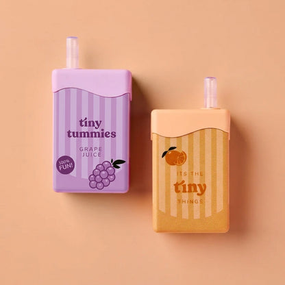 Two mini juice carton-shaped novelty items on a peach background. The left one is lavender with purple stripes, grape illustration, and the text 'tiny tummies GRAPE JUICE'.