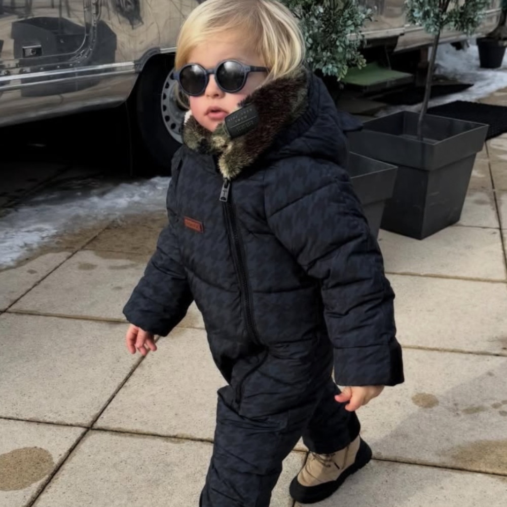 Child in a dark snowsuit with sunglasses standing on a sidewalk.