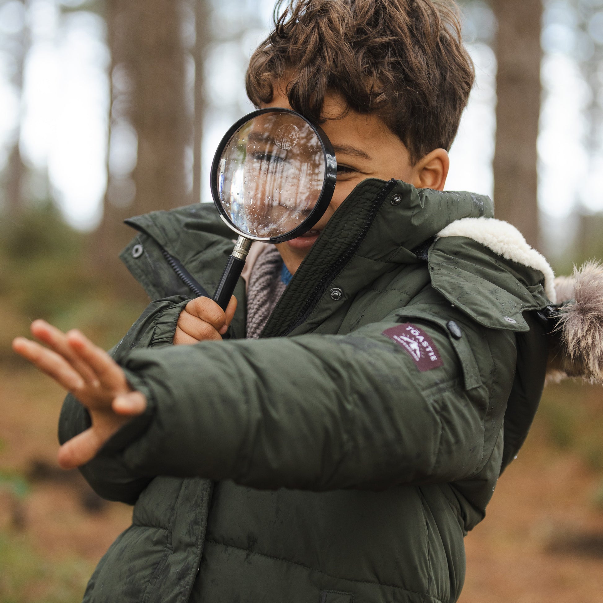 A child wearing a dark green puffer jacket with a fur-trimmed hood stands outdoors in a forest, holding a magnifying glass to one eye.