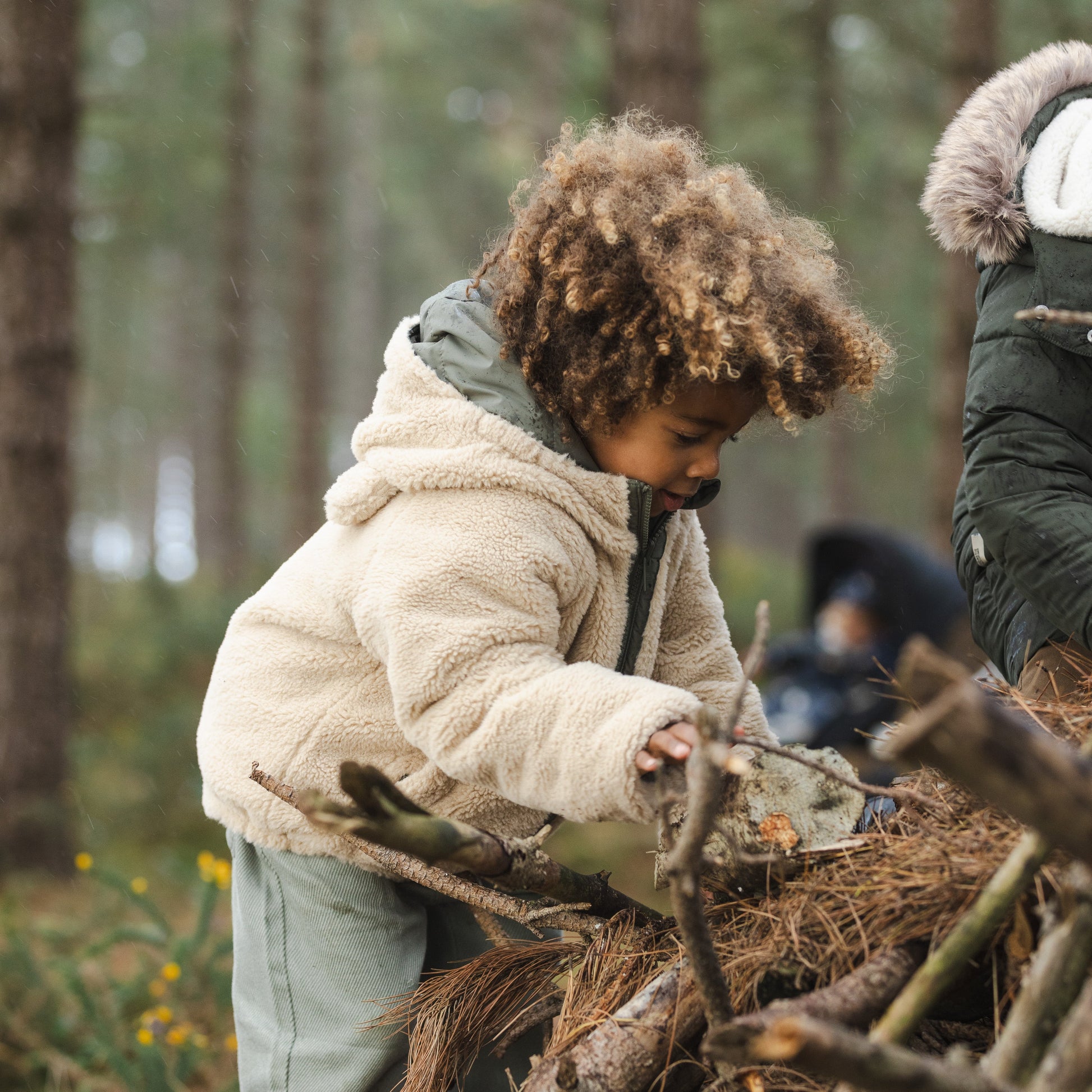 A toddler with curly brown hair wearing a cream sherpa fleece hooded jacket and gray pants is playing outdoors in a pine forest, building a twig fort with branches and pine needles. Another person is partially visible on the right.