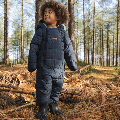A child wearing a dark navy quilted wintersnowsuit and black rubber boots standing in a pine forest on a bed of pine needles and logs.