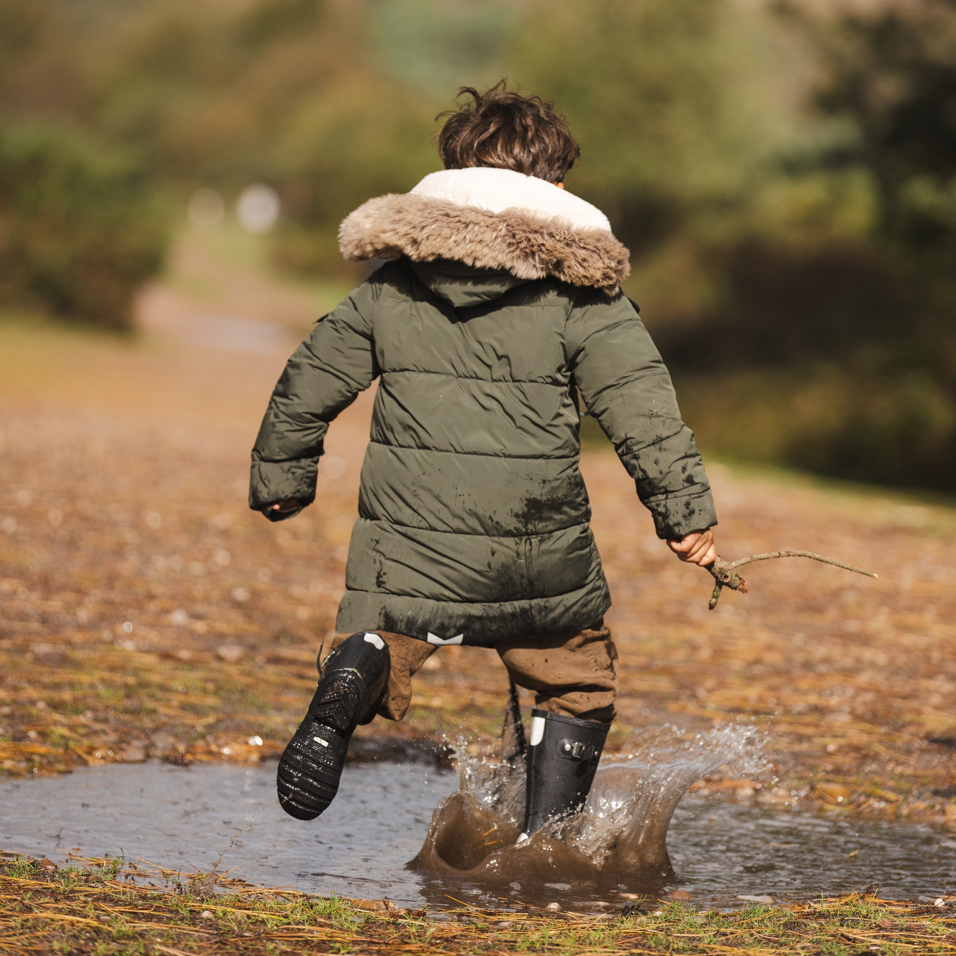 A child seen from behind running through a muddy puddle outdoors, wearing an olive green puffer parka with a beige faux-fur trimmed hood, brown trousers, and black boots, holding a small twig.