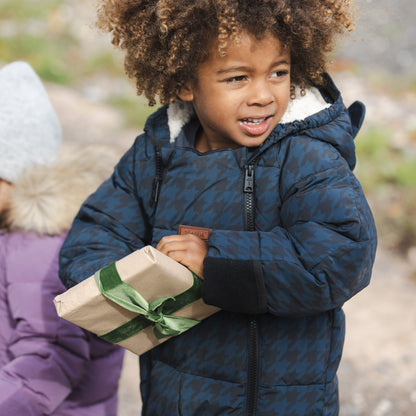 A young child with curly brown hair wearing a navy blue houndstooth patterned snowsuit and holding a wrapped gift box outdoors.