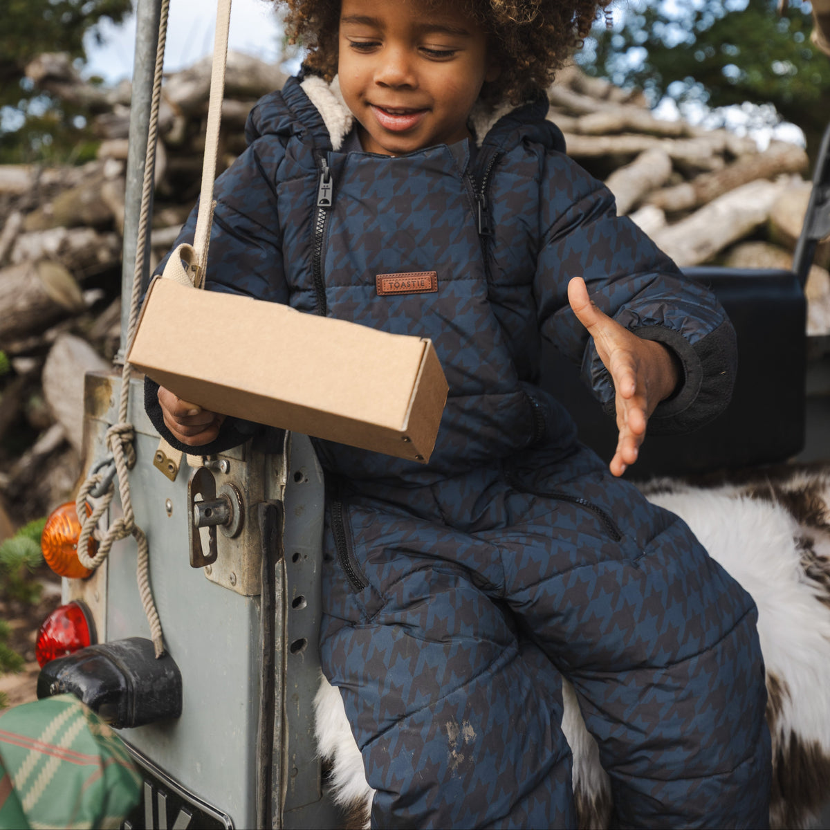 A young child with curly hair sits on the back of a vehicle outdoors, wearing a dark blue patterned snowsuit and holding a plain brown cardboard box.