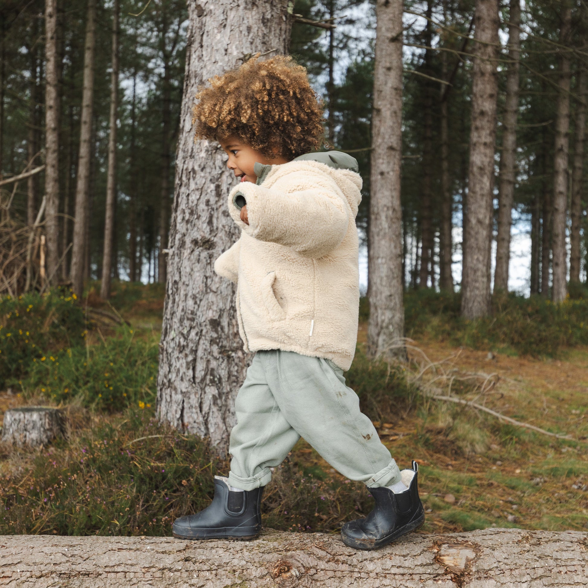 A toddler with curly brown hair walks along a fallen log in a forest, wearing a cream sherpa jacket, sage green pants, and dark rubber boots.