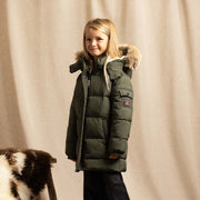 A young girl stands in a studio wearing an olive green puffer parka with a faux-fur trimmed hood, black pants, and tan boots. A patterned rug and a draped beige fabric backdrop are visible, along with a small black-and-white cowhide prop on a stool.