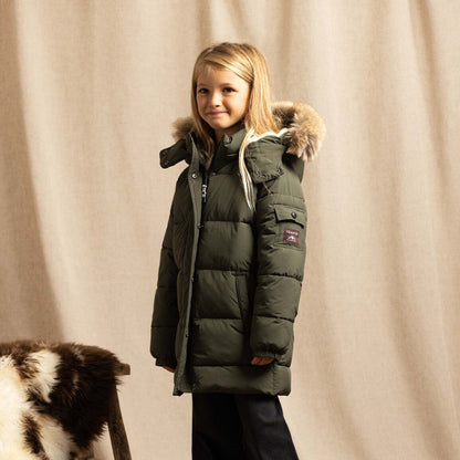A young girl stands in a studio wearing an olive green puffer parka with a faux-fur trimmed hood, black pants, and tan boots. A patterned rug and a draped beige fabric backdrop are visible, along with a small black-and-white cowhide prop on a stool.