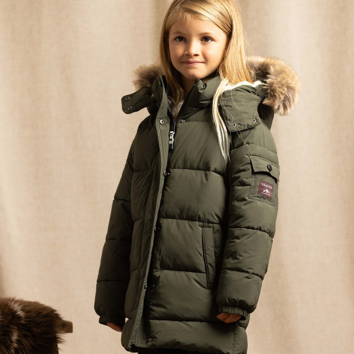 A young girl wearing a dark olive green puffer parka with a faux-fur trimmed hood, standing against a neutral beige fabric backdrop.
