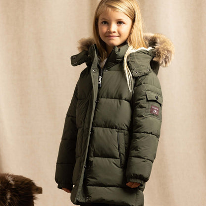 A young girl wearing a dark olive green puffer parka with a faux-fur trimmed hood, standing against a neutral beige fabric backdrop.