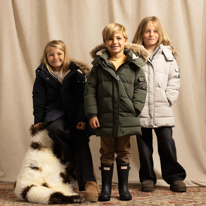 Three children wearing winter parkas pose with a black and white sheepskin rug in front of a beige fabric backdrop.