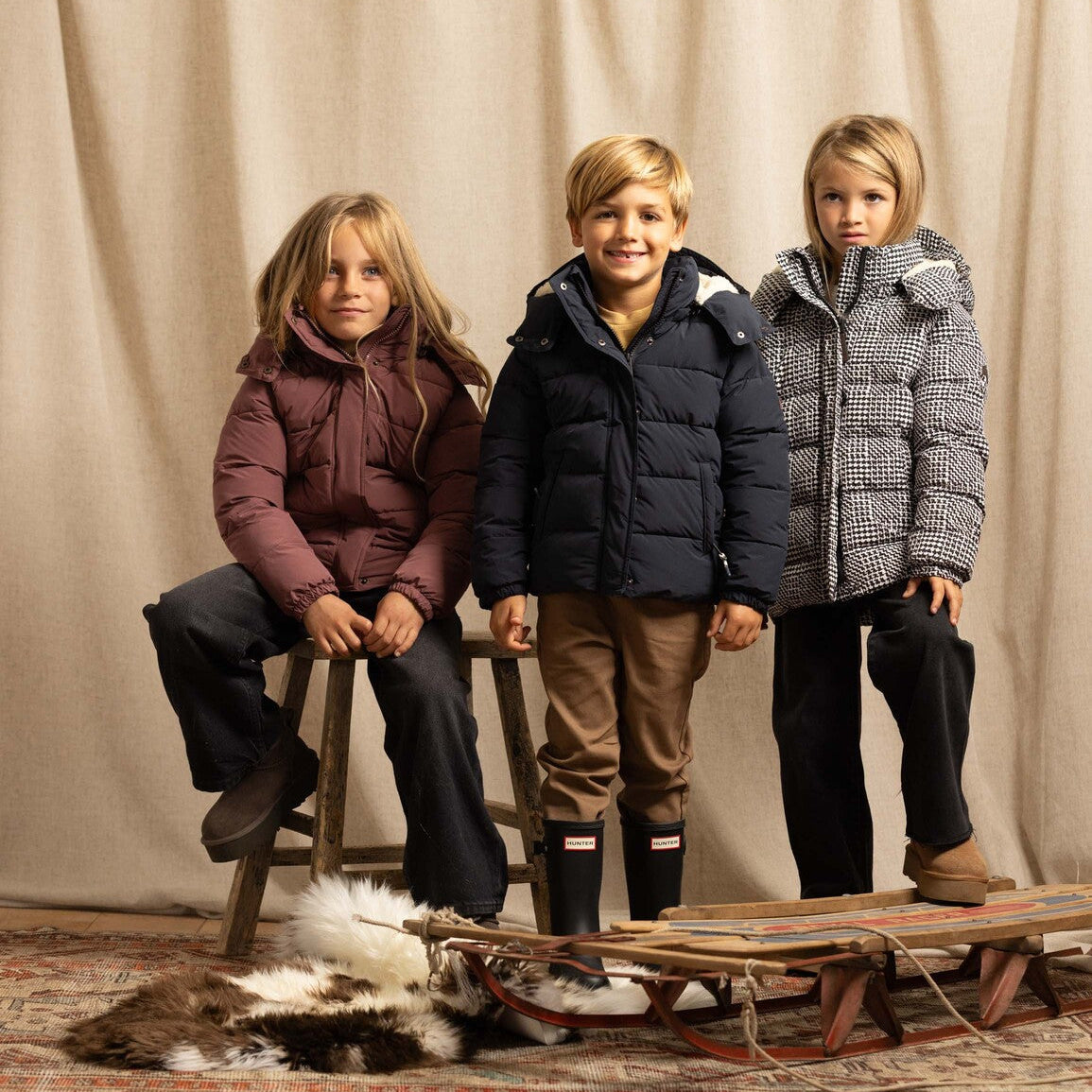Three children wearing winter jackets pose with a wooden sled and a fur rug in front of a beige curtain backdrop.