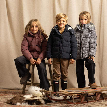 Three children wearing winter jackets pose with a wooden sled and a fur rug in front of a beige curtain backdrop.