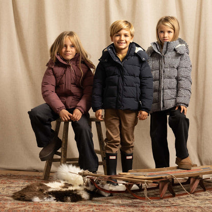 Three children wearing winter jackets pose with a wooden sled and a fur rug in front of a beige curtain backdrop.