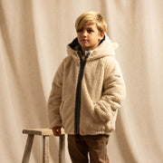 A young boy wearing a beige sherpa fleece zip-up hooded jacket, brown trousers, standing next to a rustic wooden stool in front of a draped beige fabric backdrop.