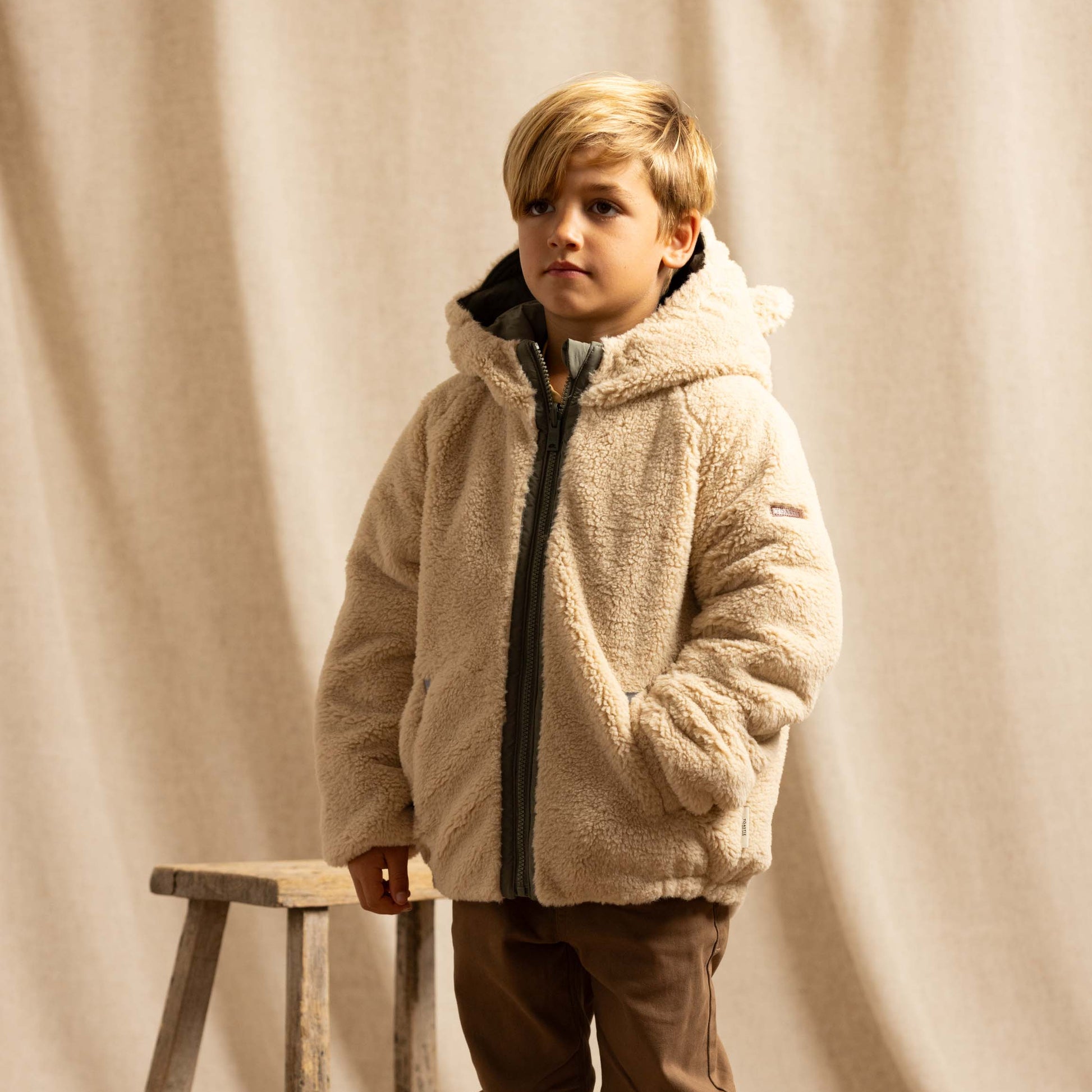 A young boy wearing a beige sherpa fleece zip-up hooded jacket, brown trousers, standing next to a rustic wooden stool in front of a draped beige fabric backdrop.