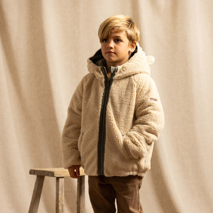 A young boy wearing a beige sherpa fleece zip-up hooded jacket, brown trousers, standing next to a rustic wooden stool in front of a draped beige fabric backdrop.