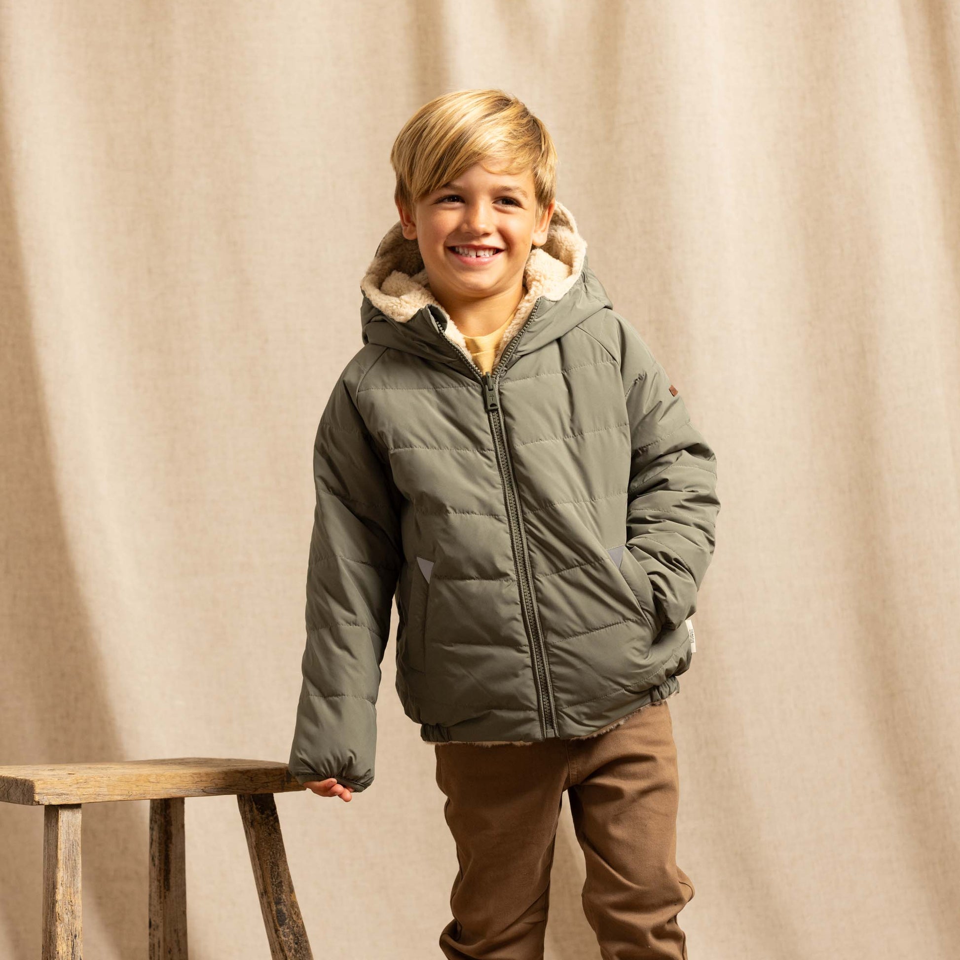 A young boy stands in a studio setting wearing an olive green puffer jacket with a beige sherpa-lined hood, brown trousers, and white sneakers. He is posed next to a rustic wooden stool in front of a draped beige fabric backdrop.