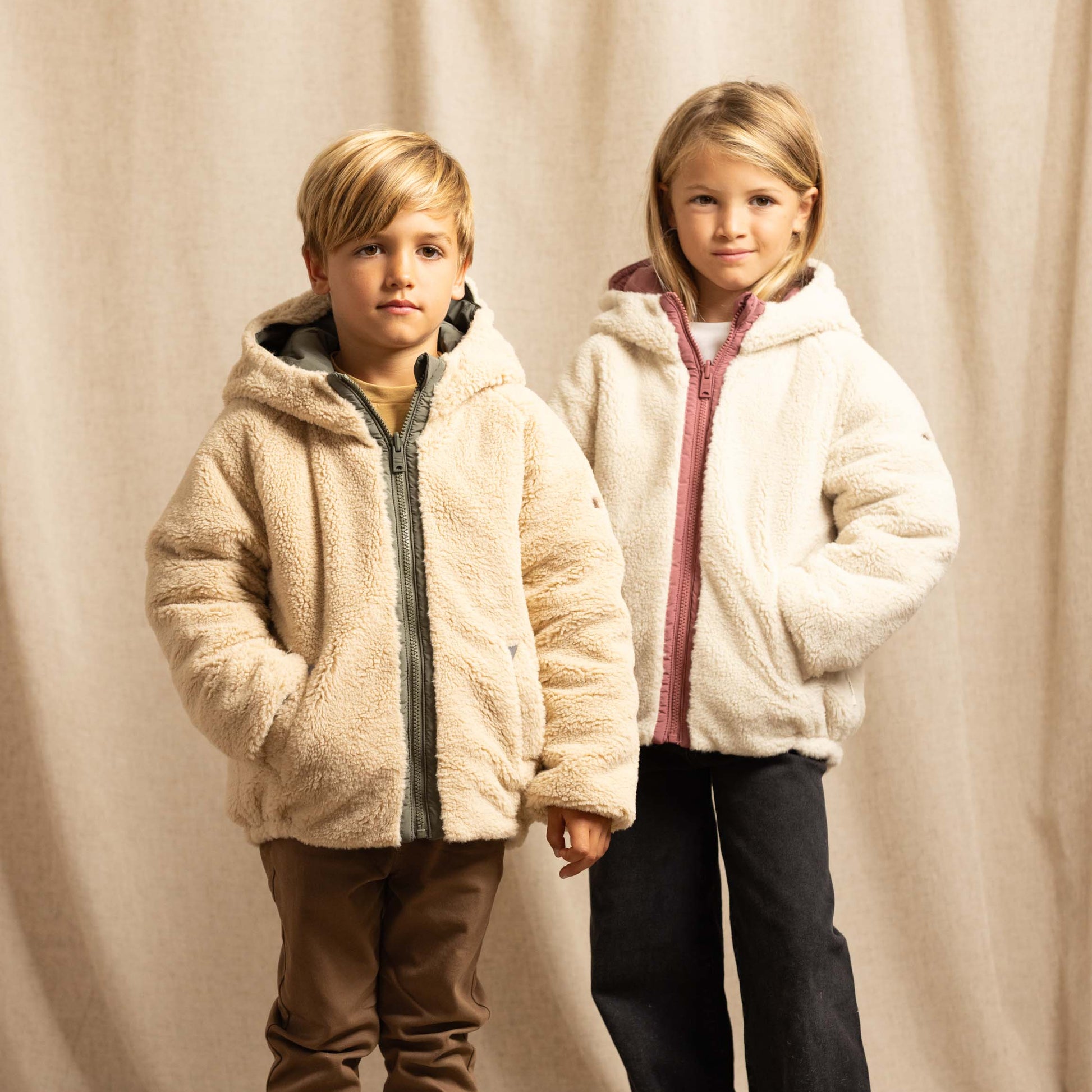 Two children standing in front of a beige fabric backdrop, wearing plush hooded jackets. The child on the left wears a cream sherpa jacket with a dark gray zipper and brown pants, while the child on the right wears a white sherpa jacket with a pink zipper and dark pants.