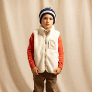 A young boy stands against a draped beige fabric backdrop wearing a cream sherpa zip-front vest over an orange long-sleeve shirt, brown pants, a navy and cream striped beanie, and white sneakers.