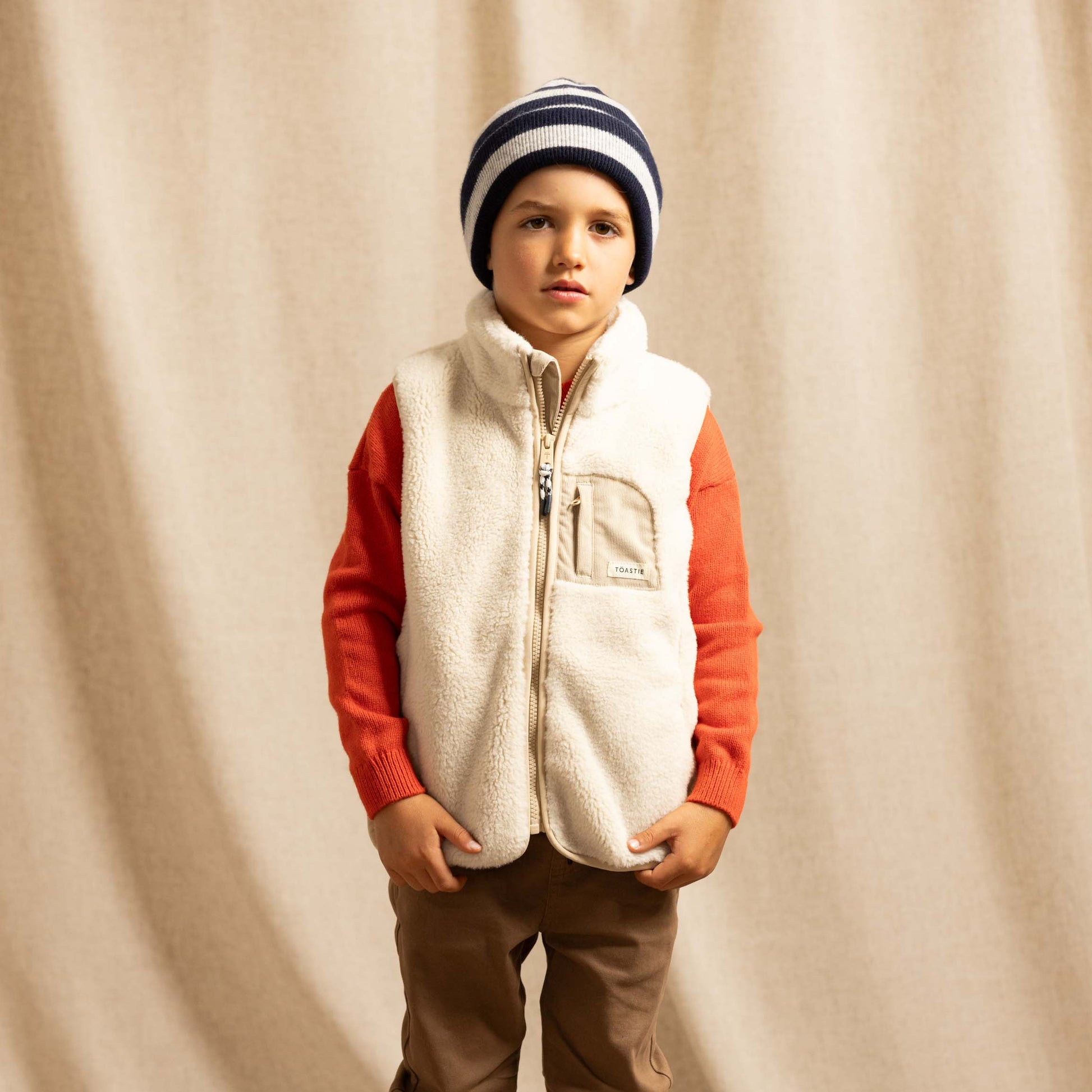 A young boy stands against a draped beige fabric backdrop wearing a cream sherpa zip-front vest over an orange long-sleeve shirt, brown pants, a navy and cream striped beanie, and white sneakers.