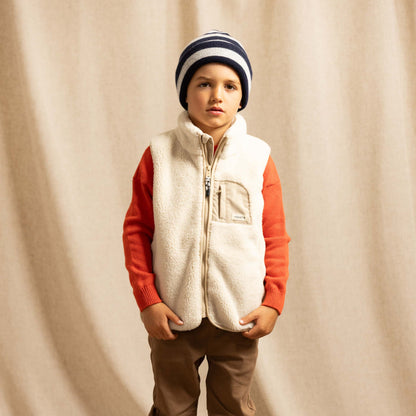 A young boy stands against a draped beige fabric backdrop wearing a cream sherpa zip-front vest over an orange long-sleeve shirt, brown pants, a navy and cream striped beanie, and white sneakers.