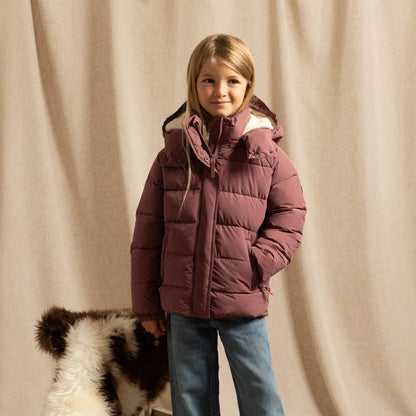 A young girl stands in front of a beige fabric backdrop wearing a dusty rose puffer jacket and blue jeans with a black-and-white cowhide rug beside her.