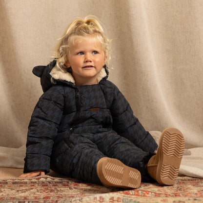 A toddler with blonde hair sits on a patterned rug wearing a dark navy quilted snowsuit with a beige sherpa-lined hood and tan boots, set against a draped beige fabric backdrop.