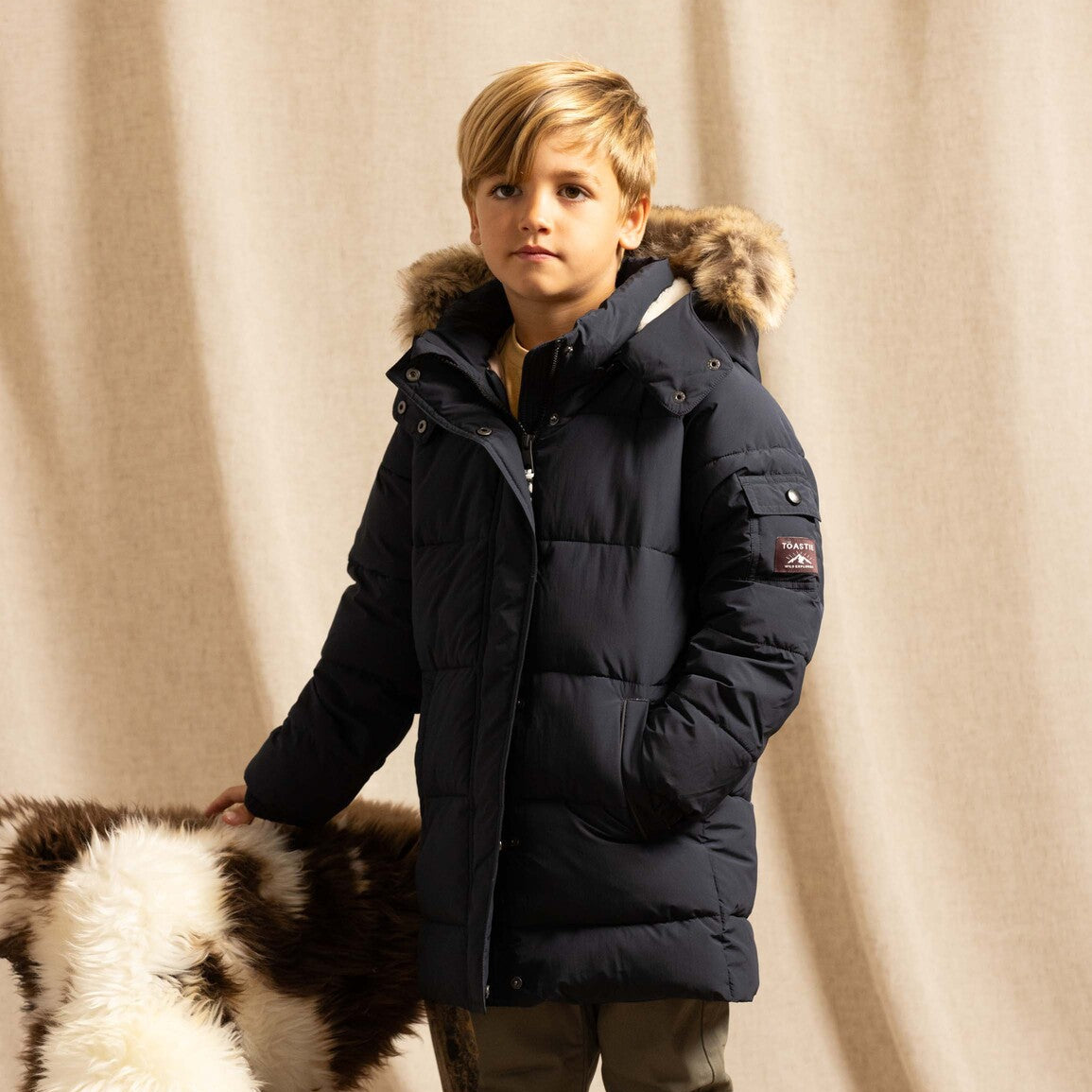 A young boy wearing a dark navy blue puffer jacket with a fur-trimmed hood, standing next to a cowhide rug in front of a beige fabric backdrop.
