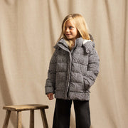 A young girl stands in a studio wearing a black and white houndstooth puffer jacket, black wide-leg pants, and brown boots, next to a rustic wooden stool against a beige fabric backdrop.