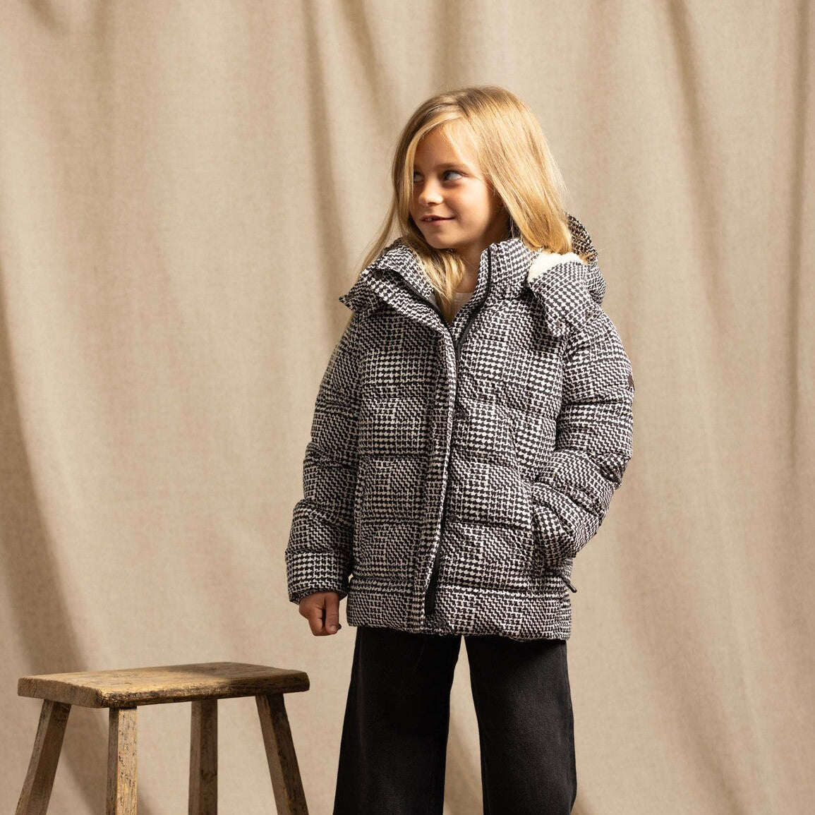 A young girl stands in a studio wearing a black and white houndstooth puffer jacket, black wide-leg pants, and brown boots, next to a rustic wooden stool against a beige fabric backdrop.