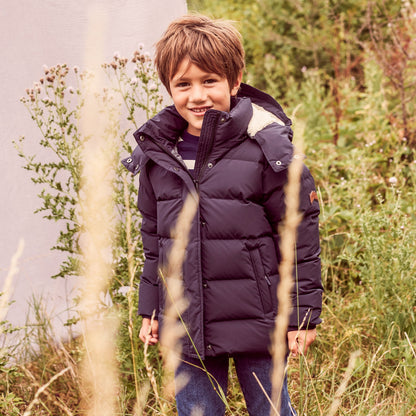 A smiling young boy wearing a dark navy blue puffer jacket with a light beige fur-trimmed hood, standing outdoors in a grassy field.