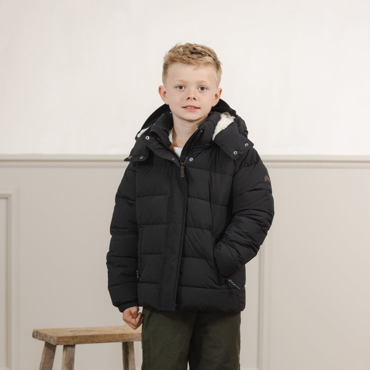 A young boy stands in a studio wearing a black puffer jacket, dark green pants, and blue sneakers, positioned next to a rustic wooden stool against a white paneled wall.