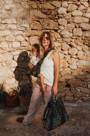 A woman and a child wearing a Wildride leopard-print baby carrier and holding a Wildride Leopard print bag, standing in front of a rustic stone wall. 