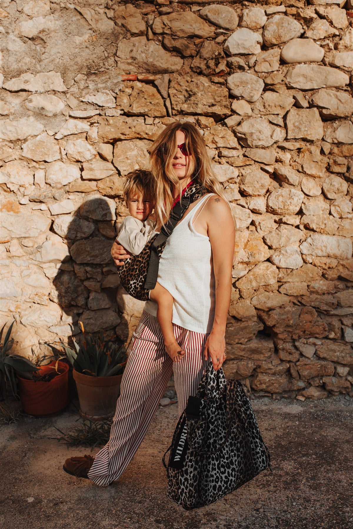 A woman and a child wearing a Wildride leopard-print baby carrier and holding a Wildride Leopard print bag, standing in front of a rustic stone wall. 