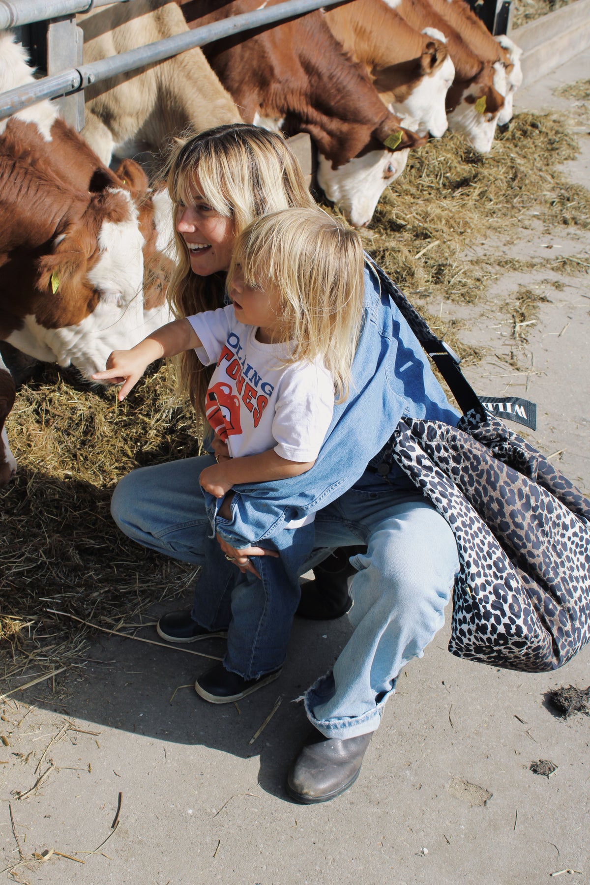 A woman and a young child crouching at a barn pen with cows, wearing a blue denim jacket and jeans, with a leopard-print shoulder bag hanging off the woman's shoulder.
