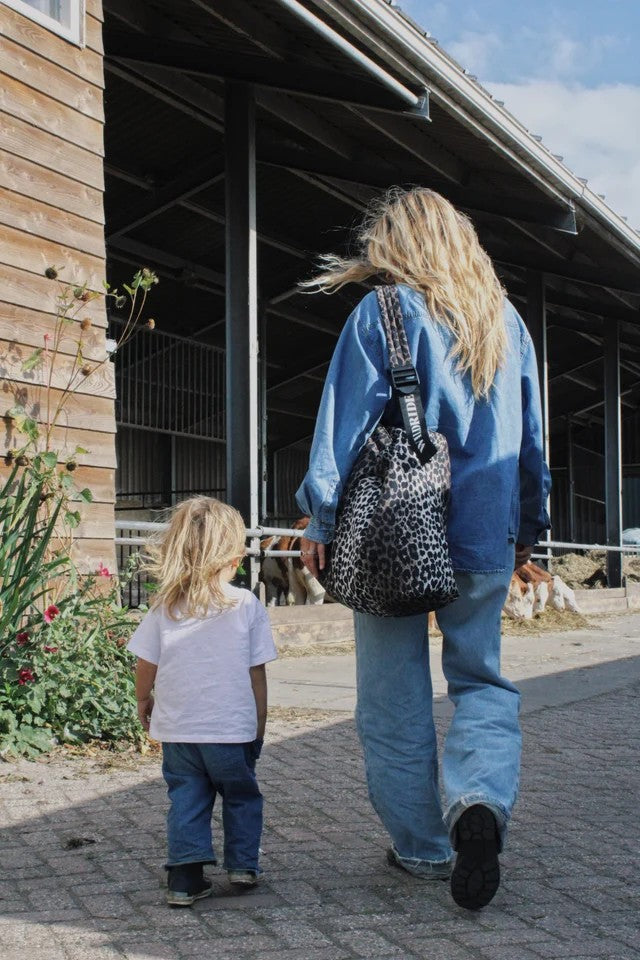 A woman and a child walk away from the camera on a paved path in front of a barn. The woman wears a blue denim jacket and jeans, carrying a black and white leopard-print shoulder bag.